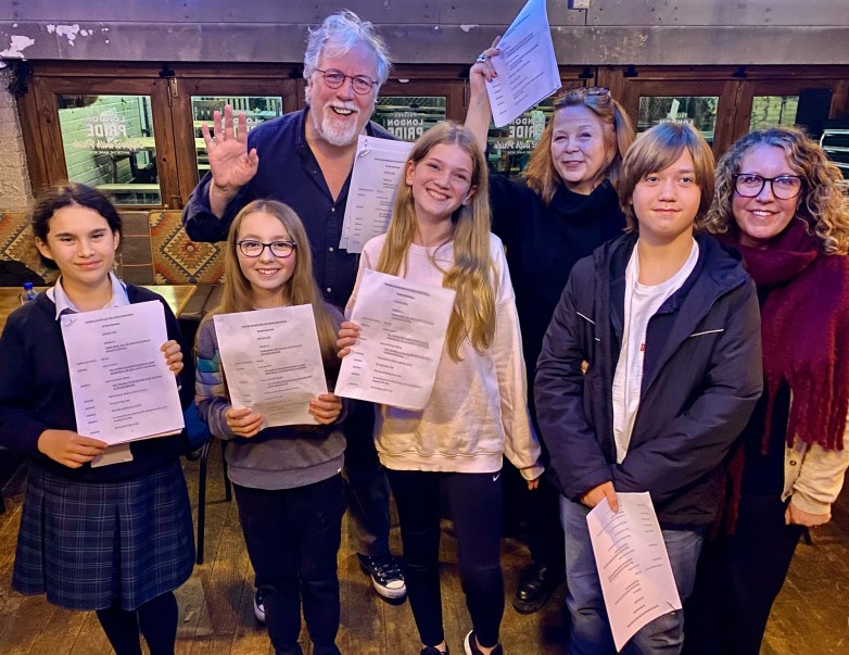 A group of six individuals, including four children and two adults, all holding papers in a warm and inviting setting, with a person in the center waving.