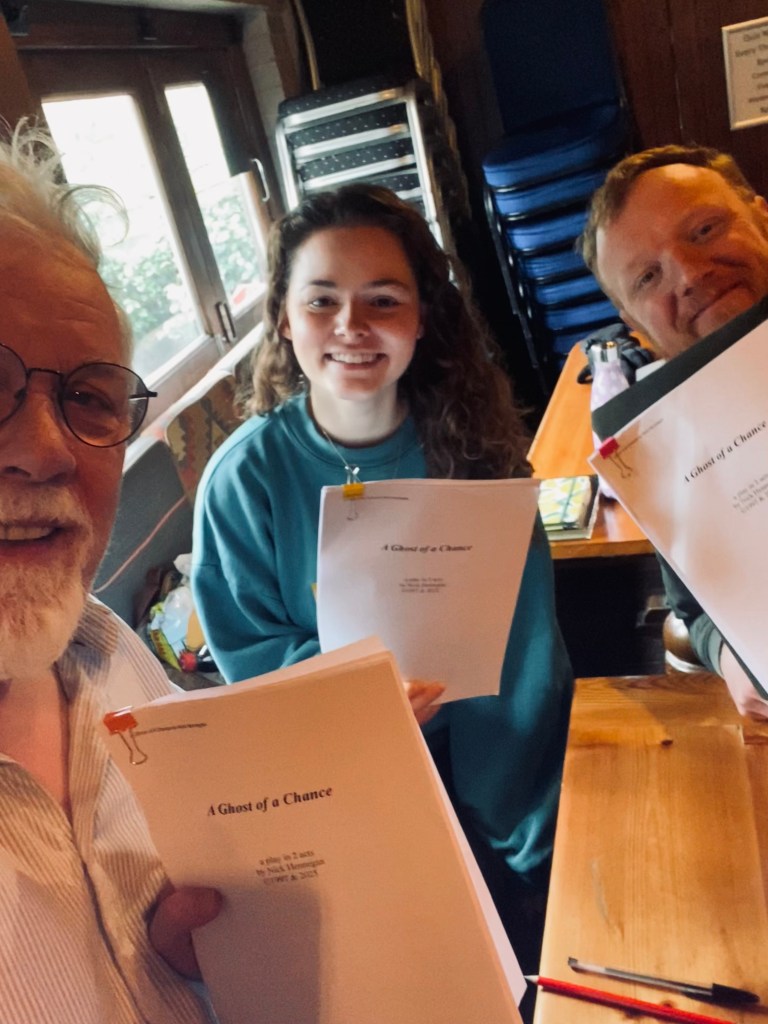 Three people smiling while holding scripts for the play 'A Ghost of a Chance' in a cozy setting.