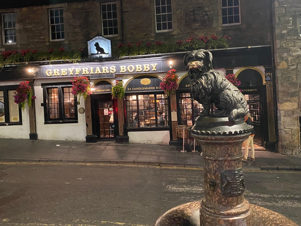 The Greyfriars Bobby pub with a statue of Greyfriars Bobby in the foreground, decorated with flowers, at night.