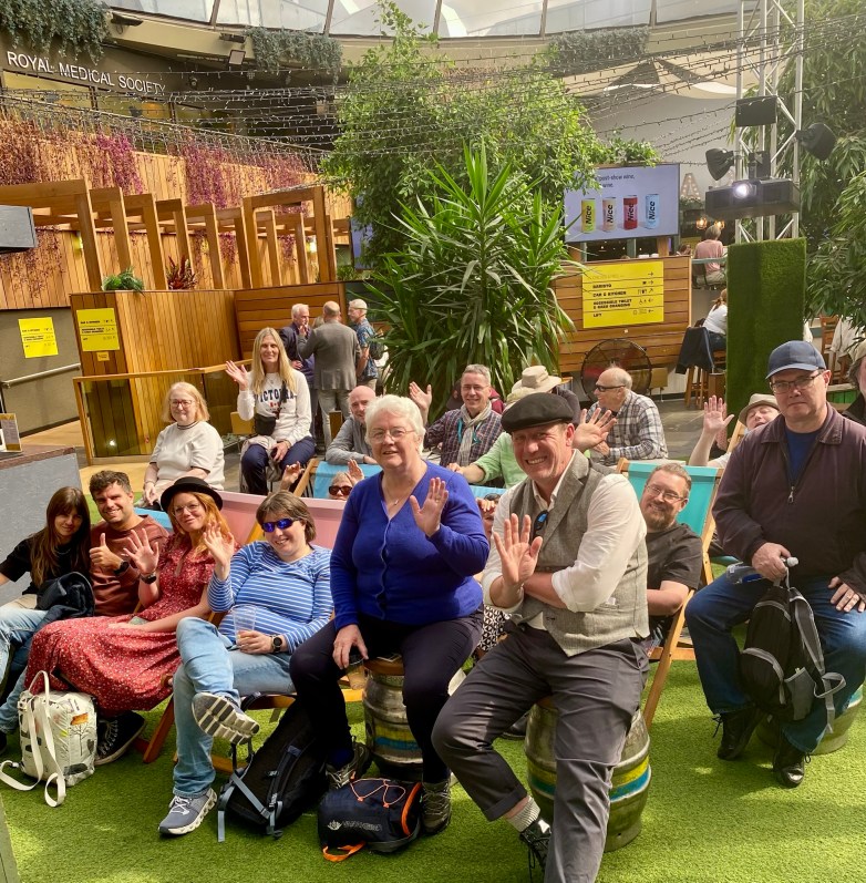 A group of people sitting in a lively indoor space, waving and smiling at the camera, surrounded by greenery and wooden decor.