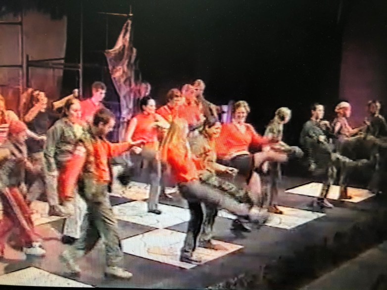 A group of performers in red and green attire engaging in a choreographed dance on stage, featuring a checkered floor.