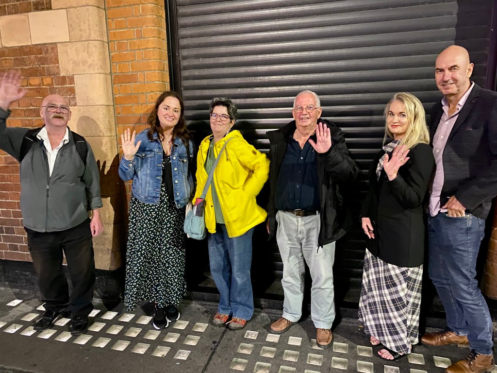 A group of six people standing outside, waving at the camera, with a brick wall and a closed shutter behind them.