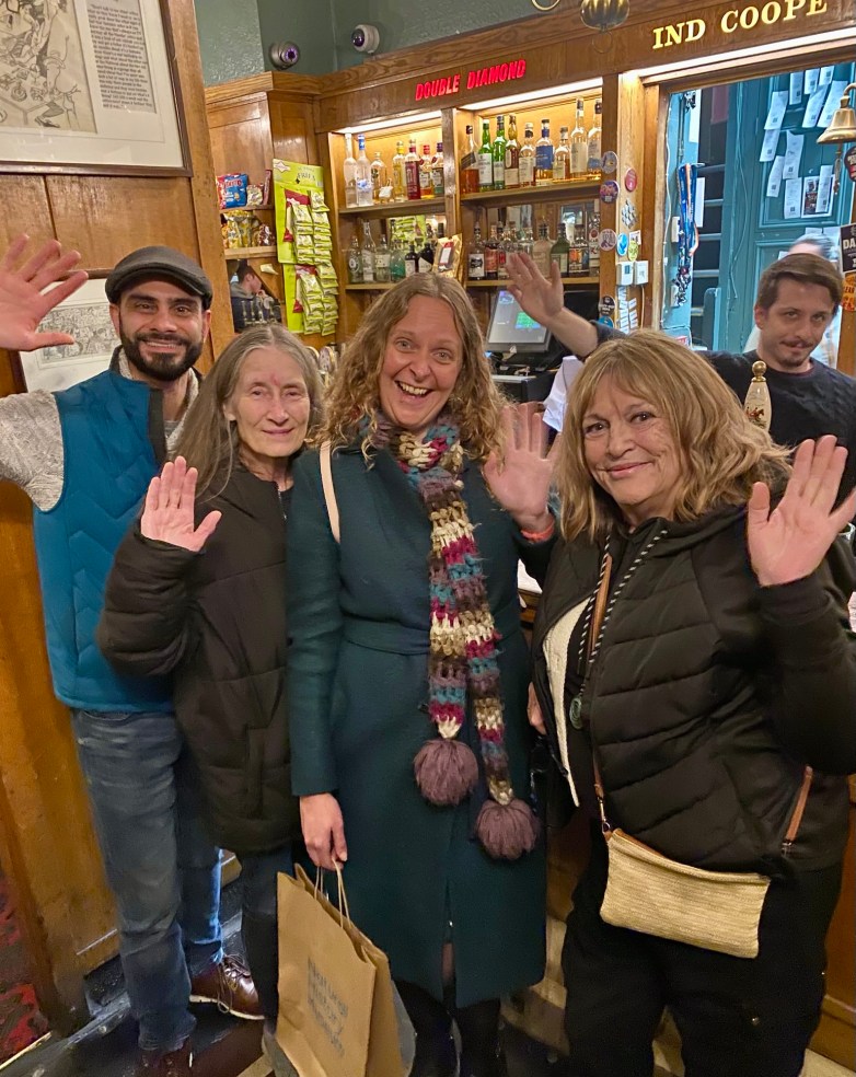 A group of five people at a pub, smiling and waving at the camera, with a bar and bottles in the background.