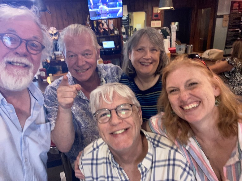 A group of five people smiling together in a casual setting, with a bar or restaurant in the background.