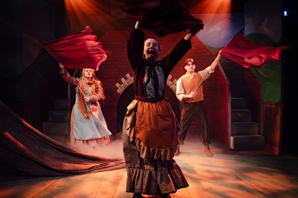 Three actors performing a scene from 'The Railway Children' on stage, with one actress waving a flag and two others in the background, illuminated by stage lights.