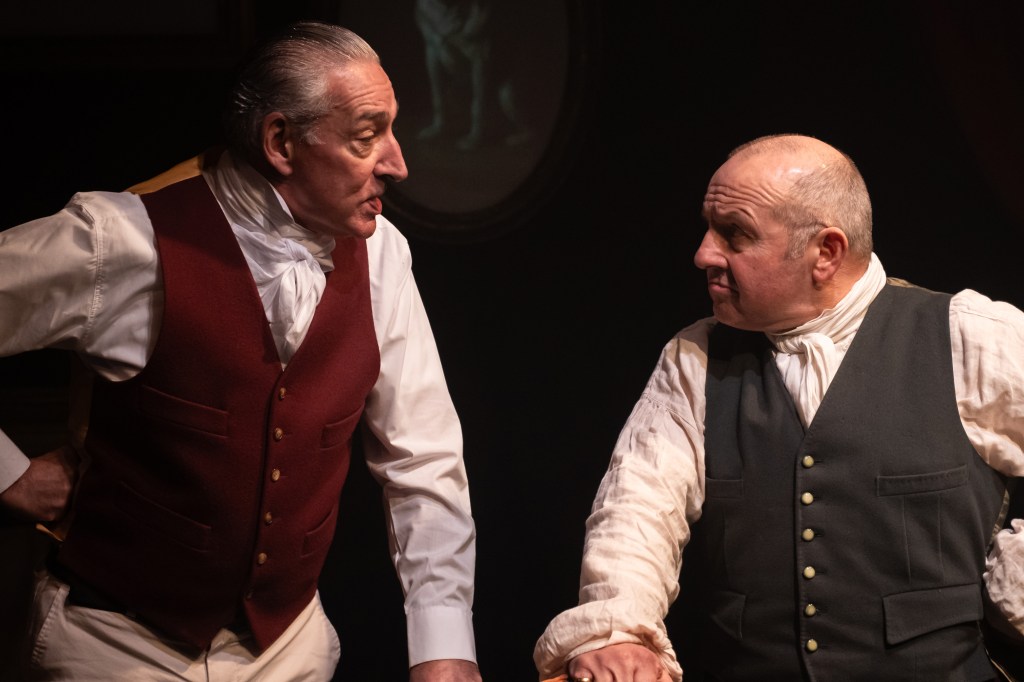 Two actors in Victorian-era costumes engaged in conversation on stage, with a dramatic backdrop.