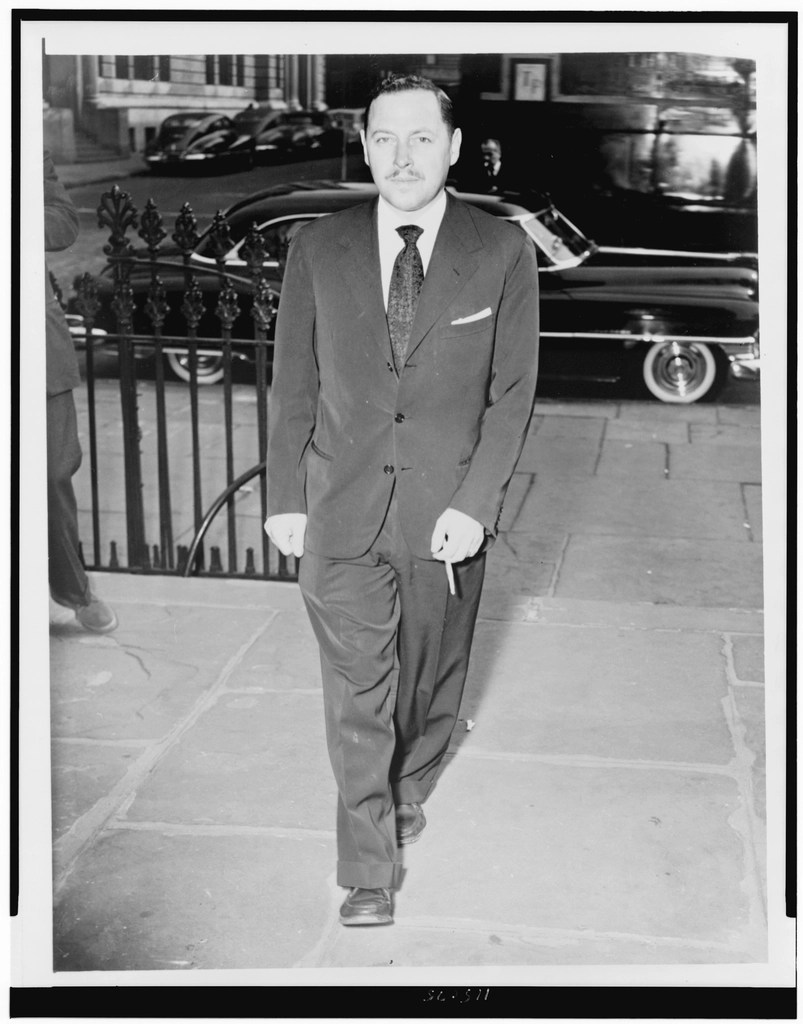 A man in a formal suit walking on a cobblestone path, holding a cigarette, with vintage cars visible in the background.