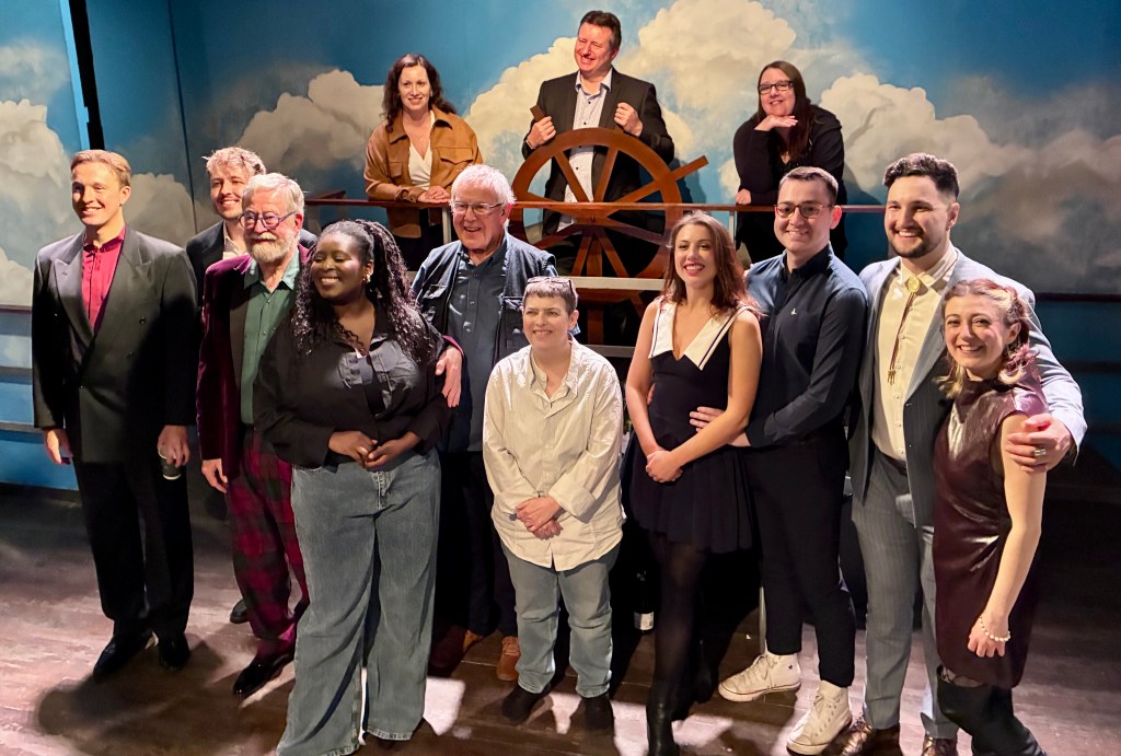 A group of diverse performers posing together on a stage, with a blue backdrop featuring clouds and a large ship's wheel. They are dressed in various styles of clothing and smiling.