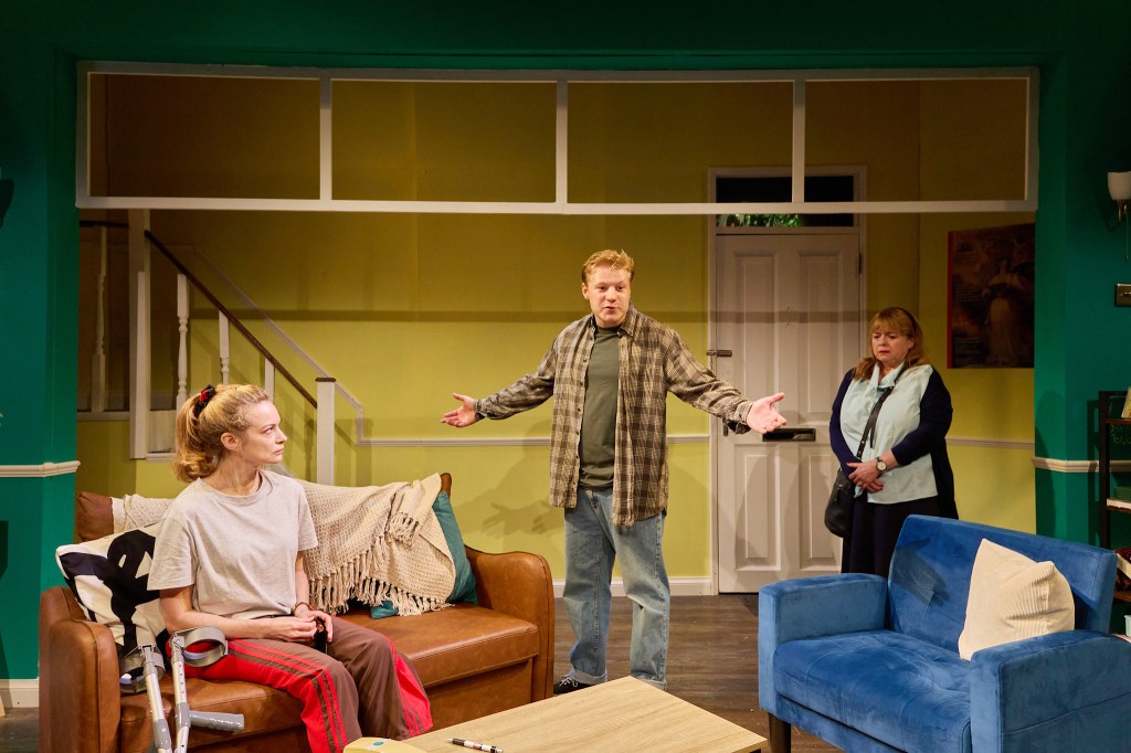 A scene from a play featuring three characters in a living room: a woman sitting on a brown couch with crutches, a man standing and gesturing, and a woman in the background observing.