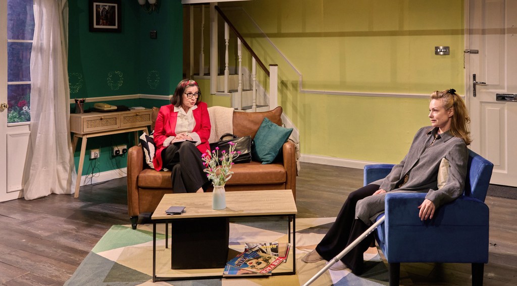 A scene from a play featuring two women seated in a living room. One woman in a red blazer is sitting on a brown couch, while the other in a gray outfit is relaxed in a blue chair, with a coffee table between them.
